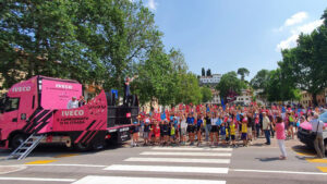 La Carovana del Giro d'Italia in Piazza Unità d'Italia a Fagagna. Un camion rosa fucsia è fermo con un intrattenitore che incita la folla a fare festa. Folla di persone e bambini che alzano le mani in alto.